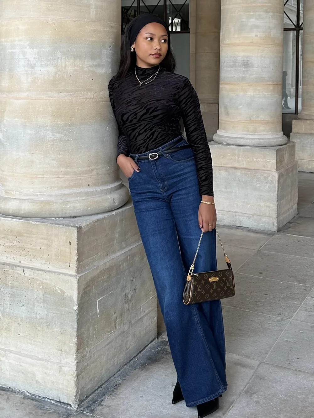 Mia in a black top and wide-leg jeans leaning on a stone column in a Paris arcade