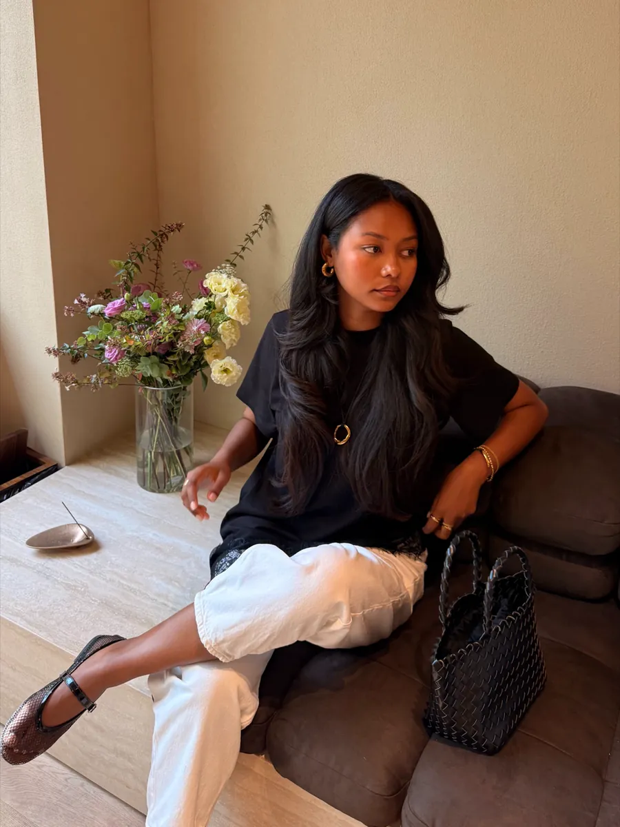 Mia seated on a sofa in a black tee, white trousers and sandals, black bag beside her