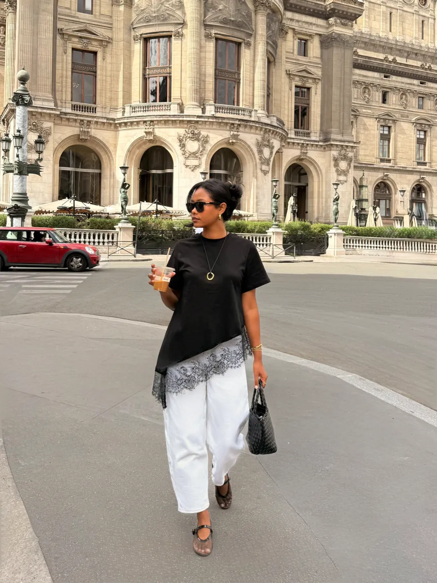 Mia walking past Palais Garnier in a black lace-trim top and white trousers, iced coffee in hand