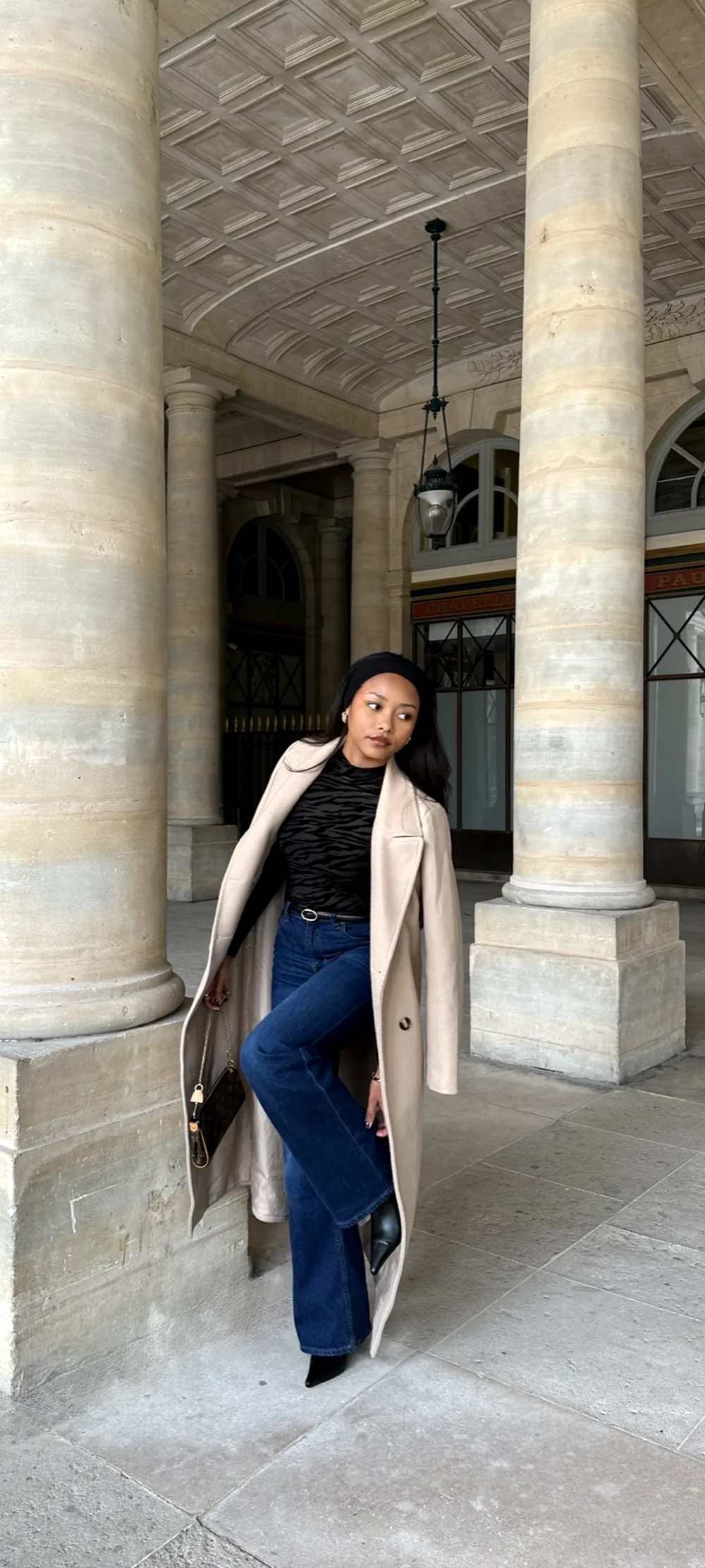 Mia leaning on a stone column in a Paris arcade, in a camel coat and flared jeans