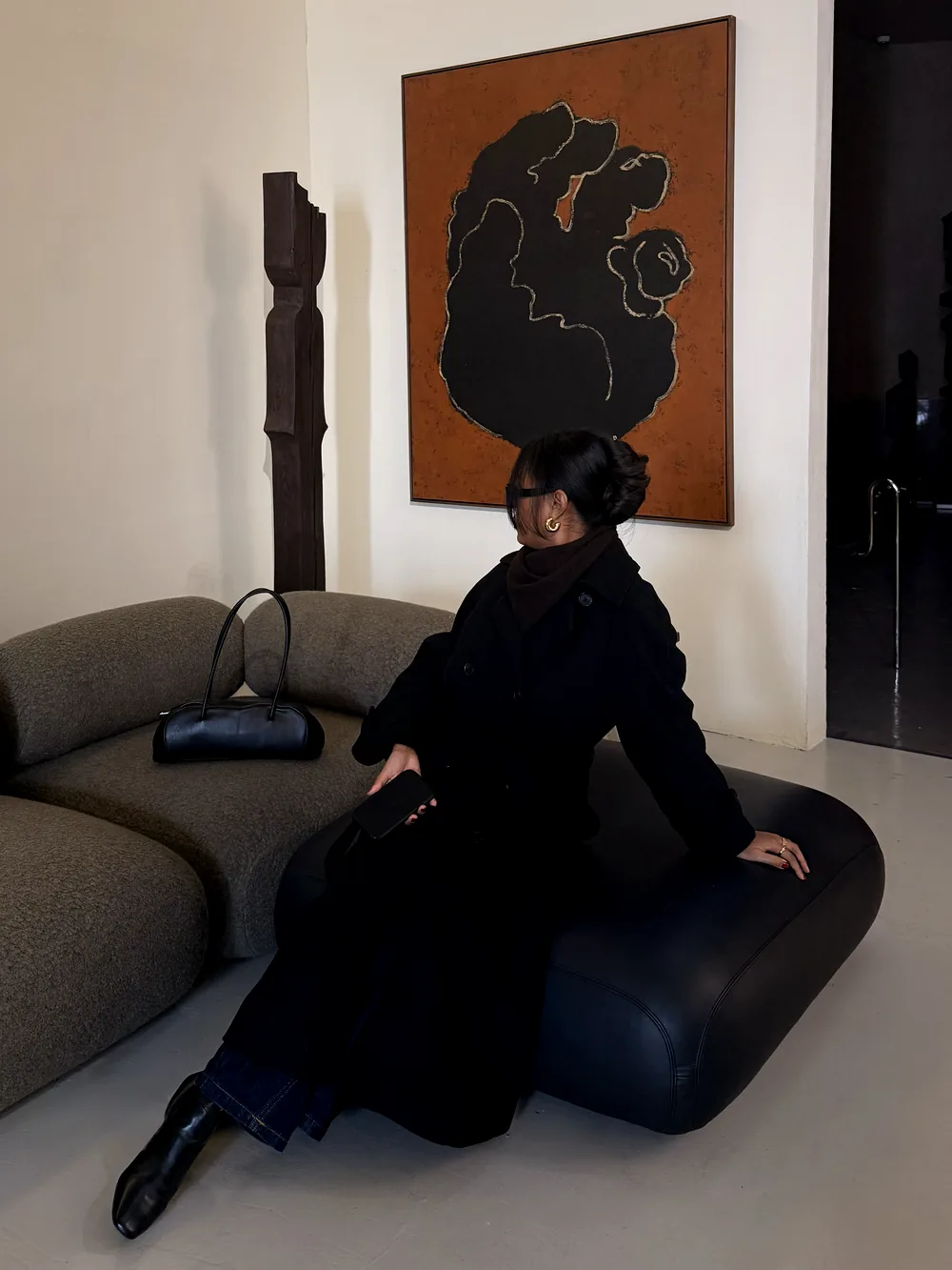 Mia in a black coat seated in a gallery lounge beside a large abstract painting