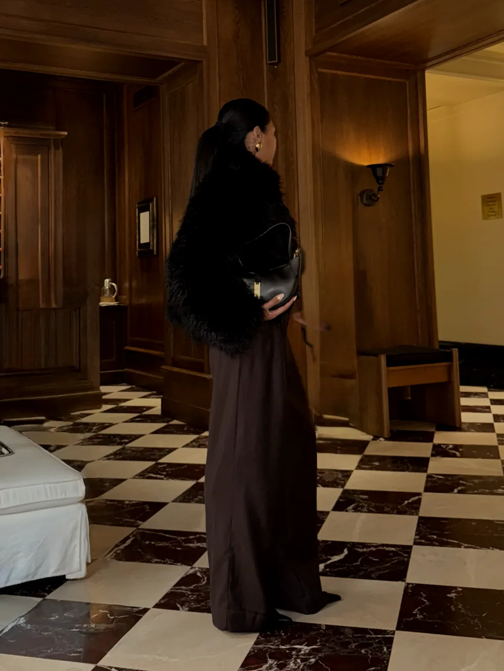 Mia in a faux-fur jacket and long skirt in a wood-panelled hotel room with a checkered floor